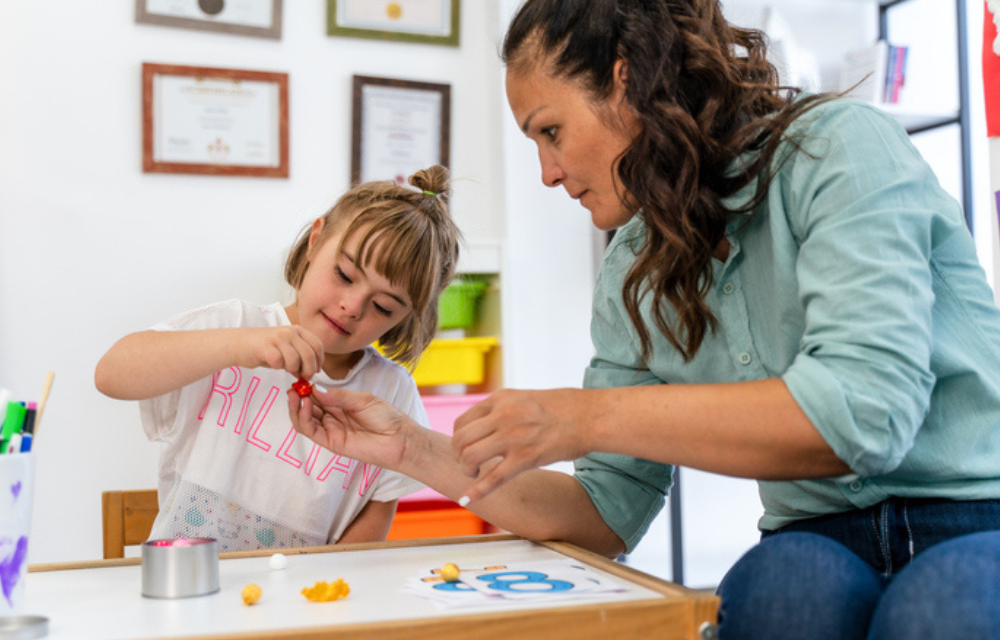 Young student is helped by teacher to play a game at a table.