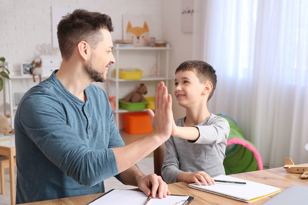 Father and son high five each other at table where both have a notebook.