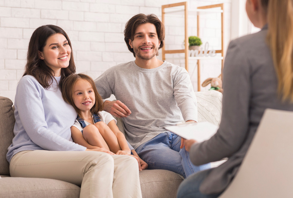 Happy couple with young daughter all sitting on a couch talking to a woman with a clipboard.