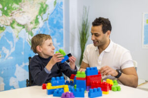 Boy in wheelchair playing with colorful blocks at table with teacher in classroom.