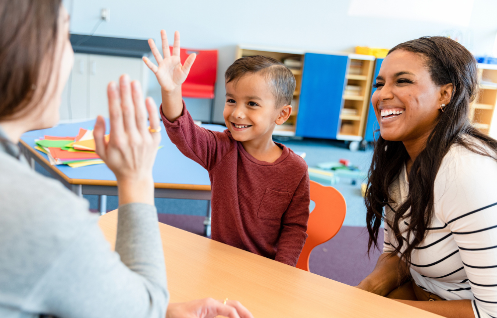 Young kid raising hand with teacher and mom at table in a classroom.