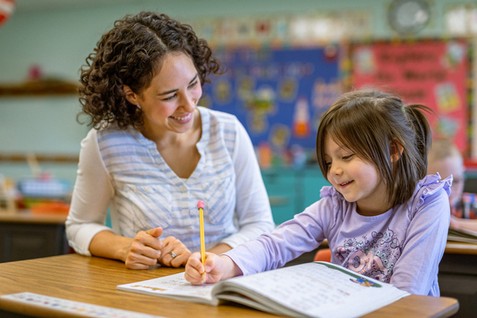 Kind teacher helps young student at desk with workbook.