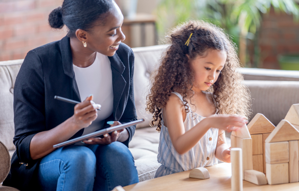 Young girl playing with blocks on table next to doctor with clipboard.
