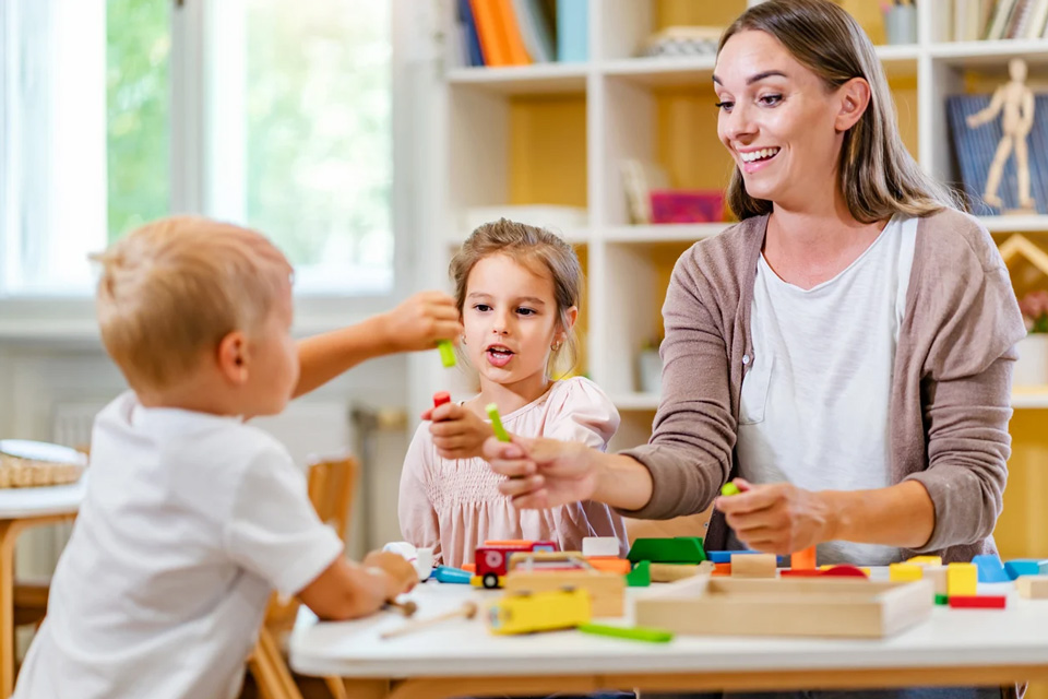 Kids and teacher play with blocks at table inside classroom.